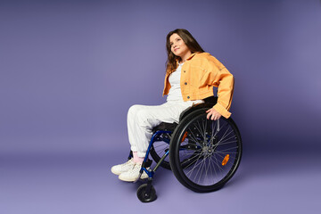 A young woman in a wheelchair sits casually on a purple studio background. She wears white pants...