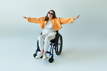 A young woman in a wheelchair wears an orange jacket and white pants, with arms outstretched in freedom and happiness