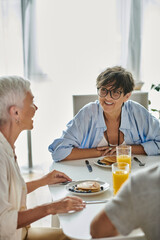 Lesbian parents enjoy a cheerful breakfast with their adult son, sharing laughter and love in their bright kitchen.
