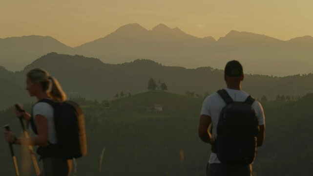 A couple with backpacks watches the sunrise over the mountains near St. Thomas Church, Slovenia