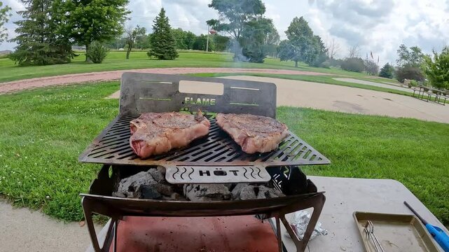 Two thick ribeye Steaks cooking on a portable, collapsible charcoal grill; concepts of tailgating, camping or picnic