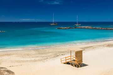 A relaxing beach view at Playa del Duque, Tenerife, with a lifeguard hut, calm turquoise waters and two sailboats