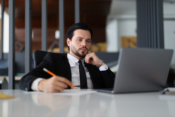 A man in a suit is sitting at a desk with a laptop and a pencil. He is deep in thought, possibly...