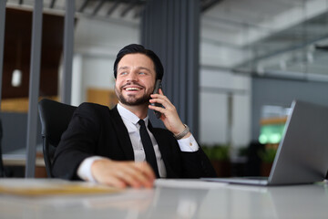 A man in a suit is talking on his cell phone while sitting at a desk. He is smiling and he is enjoying the conversation