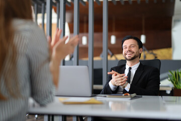 A man in a suit is sitting at a desk with a woman standing behind him. The man is smiling and...