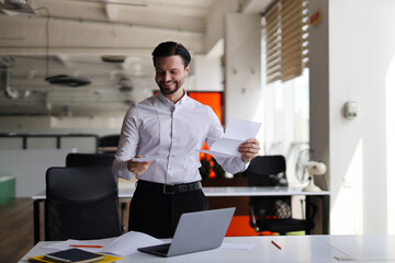 A man in a white shirt is smiling and holding a piece of paper. He is standing in front of a desk with a laptop and a chair. The scene suggests a positive and productive atmosphere