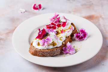 Breakfast cream cheese toast with pink culinary flowers on white plate, on pink marble background.