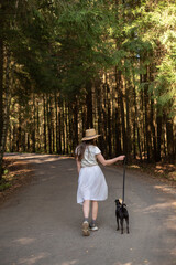 Young beautiful girl with long dark hair walking with small black dog in the forest in autumn or summer, vertical, copy space