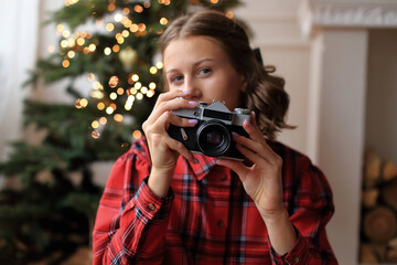 A girl in a striking red dress, holding a vintage camera in her hands. She sitting in front of a beautifully decorated Christmas tree, adorned with sparkling garlands and twinkling lights