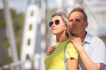 A man and an elderly woman stand next to each other. They both wear sunglasses, and the man holds the woman's hand. The scene is an expression of affection.