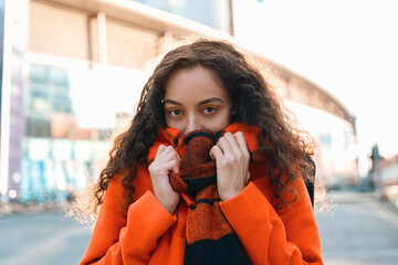 A young woman wraps herself in a cozy orange scarf while enjoying a brisk day in an urban setting