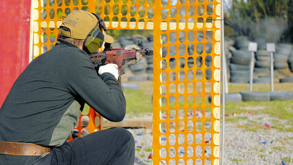 Man practicing target shooting at outdoor range. A man in protective gear aiming a firearm at a target from behind an orange barricade during a target shooting practice session.