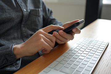 デスクの上で黒いスマートフォンを操作している手。左手で持ったスマートフォンの画面を右手の人差し指でタップしたりフリックしたりして操作している。白いキーボードも写っている。