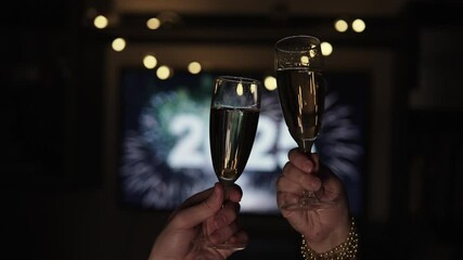 Two champagne glasses toast against blurry 2025 backdrop, suggesting celebration of New Year. Close up of clinking glasses in friendly evening setting with garlands. Romantic evening of couple.