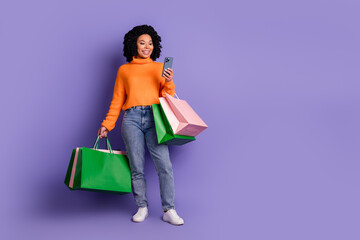 Full length photo of charming positive woman wear orange sweater holding shoppers typing modern device empty space isolated violet color background