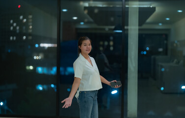 Young businesswoman is holding a smartphone while gesturing with her other hand as if presenting something to someone. The background is a blurred cityscape at night
