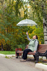 An elderly woman with an umbrella sits on a bench in a city park.