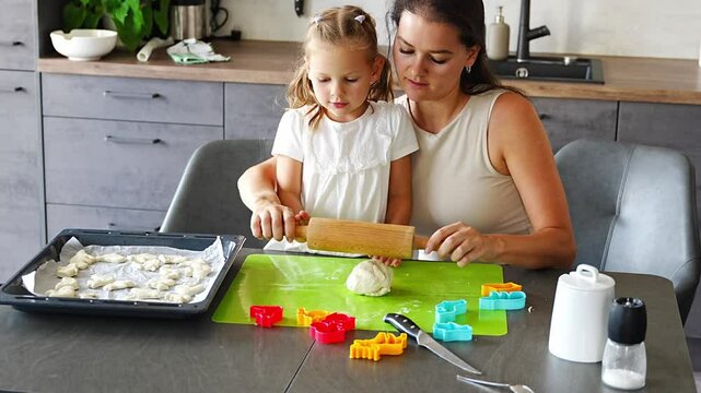 Little girl helping her mother rolling out dough with wooden rolling pinto to make cookies with dough and mold cookie cutter on home kitchen. Happy family in kitchen.