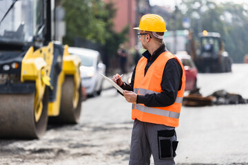 Obraz premium Construction worker inspecting roadwork during daylight, wearing safety gear on a busy urban street