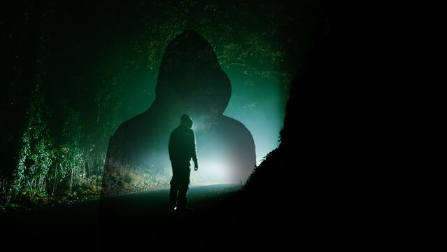 A horror concept. A ghost like hooded figure watching a man standing on a road at night. In a spooky foggy winters forest.