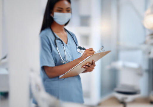 A woman in scrubs and a face mask is writing on a clipboard in a medical facility.