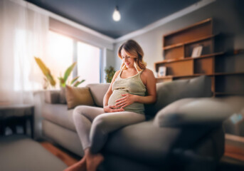 A pregnant woman sits on a couch in her living room, smiling while touching her belly.