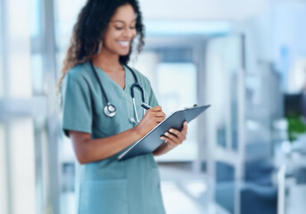 A smiling female doctor in scrubs, wearing a stethoscope, writes on a clipboard while standing in a hospital hallway.