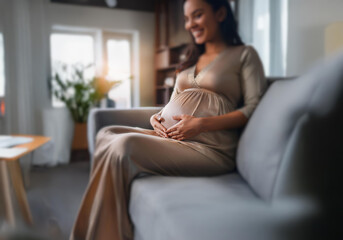 A pregnant woman in a long dress sits on a couch and cradles her belly with a gentle smile.