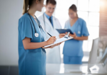 A female nurse in a blue scrub top and stethoscope writes on a clipboard with a pen. Two other medical personnel are out of focus behind her.