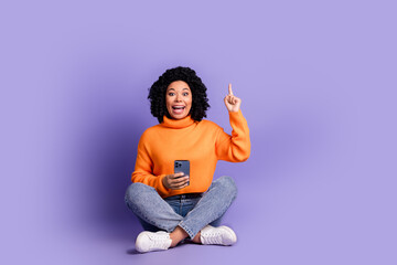 Full length photo of adorable lovely girl wear orange jumper typing modern device pointing up empty space isolated violet color background