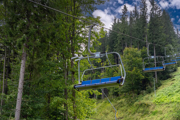 An empty lift descends to a sunny mountain resort in summer against the background of a pine...