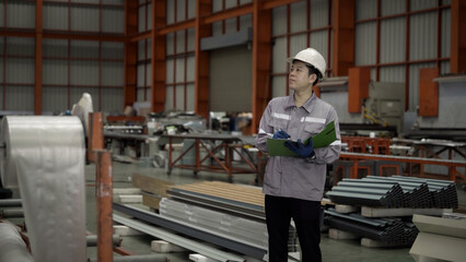 A man wearing a hard hat and safety glasses is standing in a factory with a clipboard in his hand