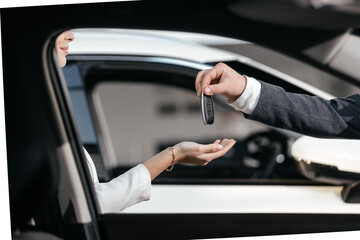 Hand holding car key remote, with modern car background. A car dealer hands over new car key to cheerful caucasian woman, indicative of a new purchase in a showroom. Rent, credit, insurance