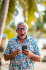 Obraz premium Senior Brazilian man in shock holding his phone, wide-eyed in a tropical beachside setting.