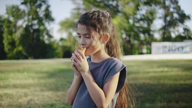 A cute little Asian girl enjoys a delicious ice cream cone on a sunny summer day in the park. Her face lights up with joy as she savors each bite, surrounded by vibrant green trees and colorful