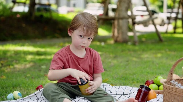 Cute Caucasian boy, about 4 years old, sit on picnic mat chill in garden, outdoor activities on sunny day, boy was covered in sweat from playing sat down rest, hand trying open cap pineapple jam jar.