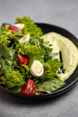 Fresh green salad with quail eggs, cherry tomatoes and guacamole in black plate on grey background