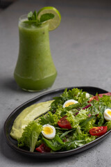 Fresh green salad with quail eggs, cherry tomatoes and guacamole in black plate and green fresh juice on grey background