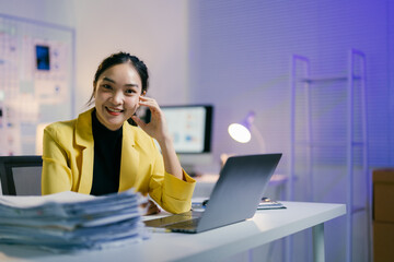 Young businesswoman works late at the office, smiling and focused on her laptop amidst paperwork. The scene captures her determination and ambition in business