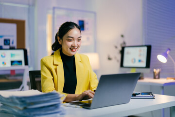 Asian businesswoman happily works late on a laptop in a modern office, surrounded by paperwork and charts, dedicated to her job