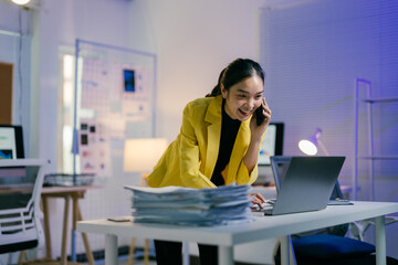 Young manager smiles while talking on her phone and working on her laptop late at night.  She is surrounded by paperwork