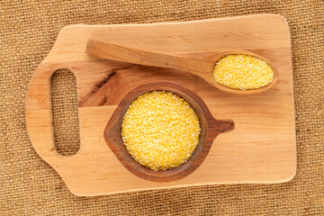 Uncooked corn grits in a wooden bowl on a jute cloth, macro, top view.