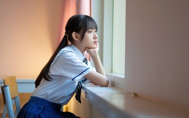 A young schoolgirl in a sailor uniform looks thoughtfully out a window.