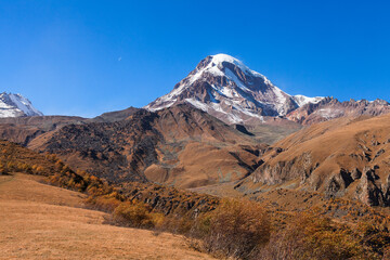 a panoramic view of autumn landscape with mountain hills surrounded by trees with yellow and red foliage at kazbegi stepantsminda in georgia near the foot of mount kazbek