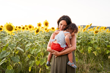 young mother and baby toddler daughter hugging and playing together outdoors in a sunflowers field in the nature. Concept of family quality time and bonding