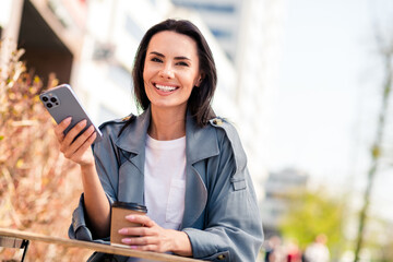 Photo of shiny attractive lady dressed grey coat smiling sitting cafe drinking tea communcating modern device outdoors town street