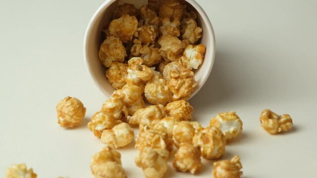 Sweet caramelized popcorn spilling out of glass, beige background, slow motion.