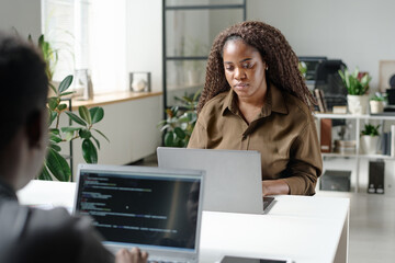 Young African American woman working in light spacious office with her unrecognizable colleague who writing code