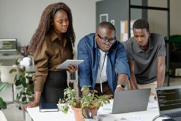 Young African American teamlead surrounded by his subordinates pointing to laptops screen and discussing written codes