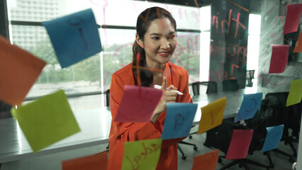 Professional young beautiful businesswoman thinking while standing at glass board with colorful sticky notes with marketing idea written. Portrait of project manager looking at mind map. Manipulator.